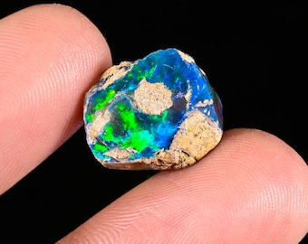 May include: A raw, unpolished opal gemstone featuring vibrant flashes of electric blue and neon green color play. The stone has an irregular, jagged shape with patches of light tan, textured matrix rock embedded across its surface.