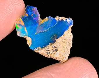 May include: A raw, irregular opal gemstone fragment featuring a vibrant, translucent blue center with flashes of green and orange iridescence. The stone has a rough, textured, beige-colored rocky matrix along its lower edge and jagged sides.