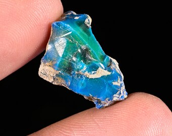 May include: A raw, unpolished gemstone fragment displaying vibrant shades of electric blue and deep emerald green. The stone has a glassy, translucent texture with irregular, jagged edges and patches of light-colored, earthy mineral matrix.