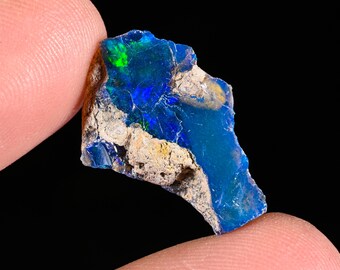 May include: A raw, unpolished opal gemstone fragment featuring a deep blue base color with vibrant green and blue flashes of light. The stone has an irregular, jagged shape with rough, textured, light brown rocky matrix material along its edges.