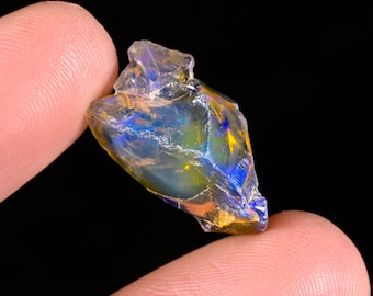 May include: A raw, translucent opal gemstone displaying vibrant flashes of blue and yellow iridescence throughout its irregular, jagged surface. The stone features a glassy texture with deep internal color play, captured against a solid black background.