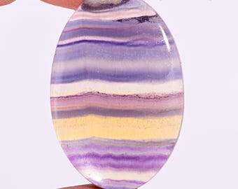 May include: An oval-shaped polished gemstone featuring horizontal bands of purple, white, and pale yellow colors. The stone has a smooth, glossy surface and is marked with the text U-1279 in the upper right corner, displaying natural mineral layering.
