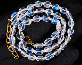 May include: A necklace featuring oval-shaped, translucent moonstone beads with a distinct blue adularescence. The beads are strung on a white cord and finished with a gold-toned metal lobster clasp and an adjustable chain link extension.