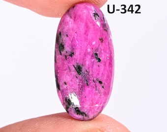 May include: An oval-shaped cabochon gemstone featuring a vibrant magenta-pink base color with scattered black mineral inclusions throughout the surface. The stone is marked with the text U-342 in the upper right corner, displaying a smooth, polished finish.