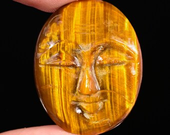 May include: An oval-shaped tiger's eye gemstone carved with a serene face featuring closed eyes, a defined nose, and a subtle mouth. The stone displays a chatoyant golden-brown color with vertical bands of light and dark brown throughout the surface.