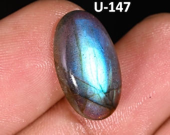 May include: An oval-shaped labradorite gemstone featuring a smooth, polished surface with iridescent blue and purple flashes. The stone displays a dark base color with visible internal fractures and the alphanumeric text U-147 in white.