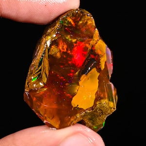 May include: A raw, unpolished fire opal gemstone with a translucent amber and deep orange base color. The stone displays vibrant flashes of red, green, and yellow light throughout its irregular, jagged surface against a dark background.