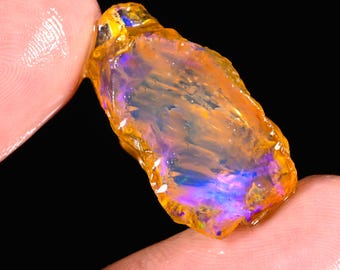 May include: A rough opal gemstone with a vibrant display of color. The stone features a mix of orange, yellow, blue, and purple hues. The opal is held between two fingers against a black background, highlighting its natural, raw form.