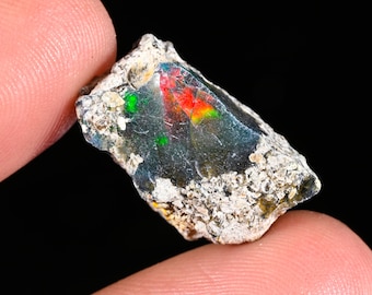 May include: A raw, unpolished black opal gemstone featuring a dark, translucent body with vibrant flashes of red and green play-of-color. The stone has a rough, textured, light-colored mineral matrix along its edges and irregular surface.