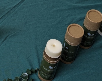 May include: Three cylindrical containers of natural deodorant are arranged on a teal surface, with a sprig of greenery in the lower left corner. The deodorant containers are made of cardboard and have a white top. The labels read "GloLillySoaps.com".