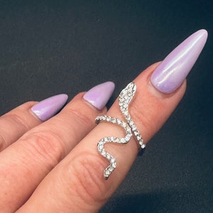 May include: A silver-toned snake ring with clear, sparkling crystals. The ring wraps around a finger, with the snake's head at the top. The ring is worn on a finger with light purple nail polish.