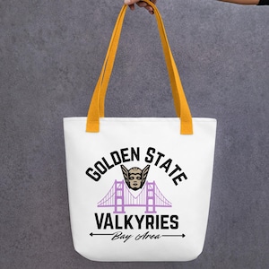 Puede incluir: Bolso tote blanco con asas amarillas. Muestra el texto "GOLDEN STATE VALKYRIES Bay Area" en negro y morado, junto con un gráfico de puente y una ilustración de Valkiria.