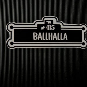 May include: A black and white sign displaying the word "BALLHALLA" and the number "415". The sign features a decorative border and a bear icon. The background is black with vertical lines.