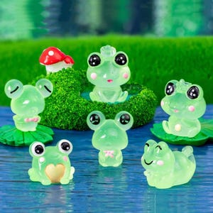 May include: A selection of translucent green frog figurines with black eyes and pink cheeks. Some frogs are on lily pads, others are standing or sitting. A red and white mushroom is in the background.