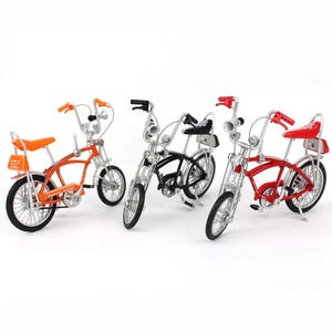May include: Three miniature bicycles in orange, black, and red. Each bike has a chrome frame, high handlebars, and a small seat. The orange bike has an orange seat and a small bag on the back. The black and red bikes also have a small seat.