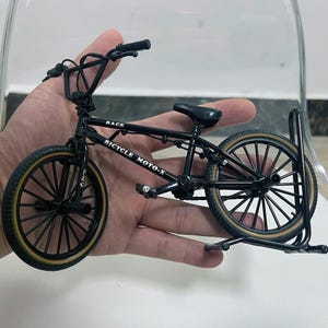 Könnte beinhalten: Ein kleines schwarzes BMX-Fahrradmodell mit dem Text "RACE BICYCLE MOTO-X" auf dem Rahmen. Das Fahrrad hat schwarze Reifen mit beigefarbenen Seitenwänden, einen schwarzen Sattel und einen Lenker. Es wird auf einer transparenten Oberfläche präsentiert.