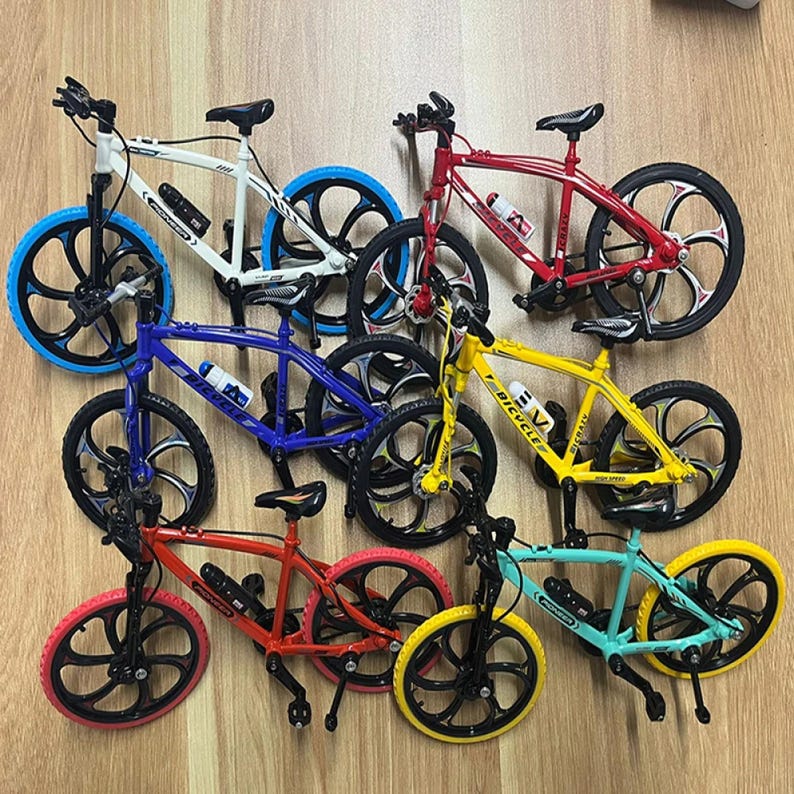 Op de afbeelding: Een verzameling miniatuurfietsen in verschillende kleuren, waaronder wit, rood, blauw, geel en turquoise. Elke fiets heeft zwarte wielen met een uniek spaakontwerp en een kleine waterfles. De fietsen zijn gerangschikt op een lichtbruin oppervlak.