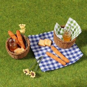 May include: A miniature picnic scene on green grass. Two woven baskets hold bread, toast, and a jar of honey. A blue and white checkered cloth and decorative flowers complete the setting.