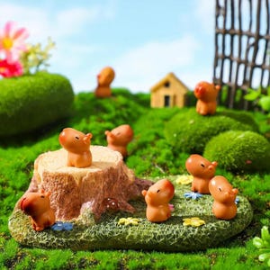 Könnte beinhalten: Eine Miniaturgartenszene mit mehreren kleinen, orangefarbenen Capybara-Figuren. Die Szene umfasst einen Baumstumpf, grünes Moos, Miniaturblumen und ein kleines Haus, alles vor dem Hintergrund von grünem Gras und blauem Himmel. Die Figuren sind der Fokus.