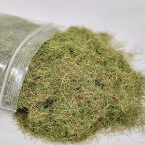 Może przedstawiać: Przezroczysta plastikowa torba, częściowo otwarta, wysypująca stos sztucznej trawy. Trawa jest mieszanką zieleni i brązu, o teksturowanym wyglądzie. Produkt jest prawdopodobnie używany do modelarstwa lub rękodzieła.