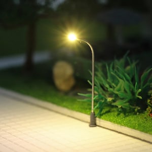 May include: Miniature street lamp with a bright yellow light. The lamp post is silver and has a curved arm. The base is dark gray. The lamp is set on a miniature sidewalk with green grass and bushes.