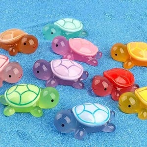 May include: A selection of small, brightly coloured turtle ornaments in shades of orange, pink, purple, green, yellow, and blue. Each turtle features a detailed shell pattern and a smiling face. The turtles are displayed on a textured, blue surface.