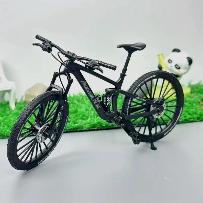 Może przedstawiać: Czarny model roweru g&oacute;rskiego z napisem "MODEBIKE CYCLE" na ramie. Rower ma czarne opony, czarne siodełko i czarną kierownicę. Rower stoi na szarej powierzchni z figurką pandy w tle.