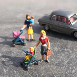 Puede incluir: Figuras en miniatura de madres con niños y cochecitos, y un coche gris. Una madre sostiene a un bebé, otra empuja un cochecito rosa y una tercera está de pie con un niño en un cochecito azul. Las figuras están pintadas con colores brillantes.