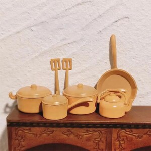 May include: A collection of miniature kitchen cookware in a warm, light brown color. The set includes pots, pans, a kettle, and cooking utensils. The items are arranged on a wooden surface, suggesting a toy or decorative theme.