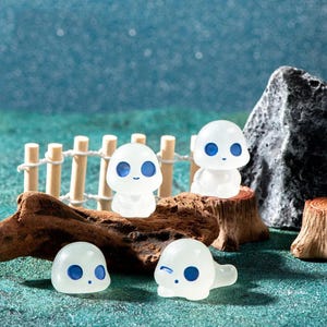 May include: Several small, white, ghost-shaped figurines with blue eyes and smiling expressions. Some are standing, others lying down. They are displayed on a wooden base, with a miniature fence and a rock in the background, creating a whimsical scene.