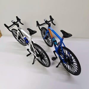 Könnte beinhalten: Zwei Rennräder, eines weiß und eines blau, werden gezeigt. Das weiße Fahrrad hat schwarze Räder und eine blaue Wasserflasche. Das blaue Fahrrad hat orangefarbene Akzente und eine blaue Wasserflasche. Beide haben schwarze Sitze und Lenker.