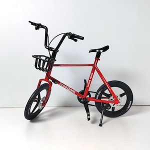 May include: A red miniature bicycle with black handlebars, seat, and tires. The bike has a small basket in the front and the word "Freedom" printed on the frame. The rear wheel has the text "EXPLORE" and "ALWAYS YOUNG".