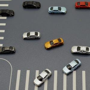 May include: A selection of miniature toy cars in various colours, including black, silver, gold, red, and blue, arranged on a grey surface with white road markings. The cars are small, detailed replicas of modern vehicles.