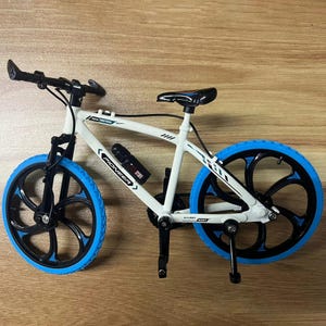 Op de afbeelding: Een kleine witte fiets met blauwe en zwarte details. De fiets heeft blauwe banden, een zwart stuur en een zwart zadel. Op het frame staat het woord "PIONEER" en er is een waterfles aan het frame bevestigd.