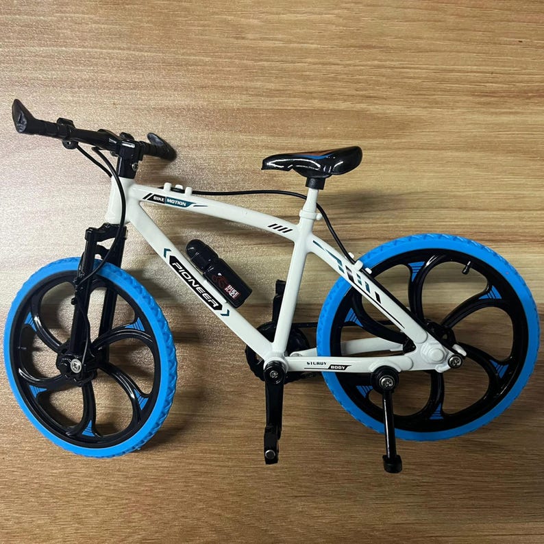 Op de afbeelding: Een kleine witte fiets met blauwe en zwarte details. De fiets heeft blauwe banden, een zwart stuur en een zwart zadel. Op het frame staat het woord "PIONEER" en er is een waterfles aan het frame bevestigd.