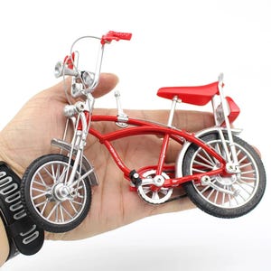 May include: A miniature red bicycle with silver accents, held in a hand. The bike features a curved frame, a red seat, and silver fenders. The tires have a black and silver design. The handlebars and front lights are silver.
