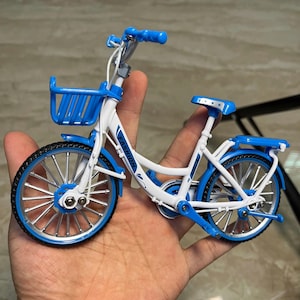 May include: A miniature bicycle with a white frame and blue accents. The bike features a front basket, fenders, and a rear rack. The tires are black with silver spokes. The word "FASHION" is printed on the frame.