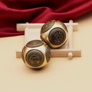 May include: Two gold-coloured, spherical decorative objects with intricate patterns and Chinese characters. The objects are resting on a small, light-coloured wooden tray, set against a backdrop of a deep red fabric. The spheres have a vintage, antique aesthetic.