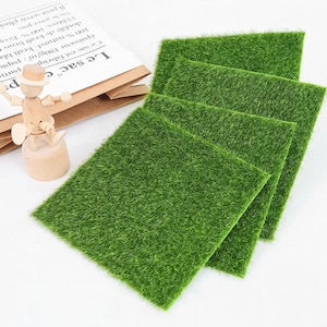 May include: Four square artificial grass mats in a bright green color. The mats are arranged in a slightly overlapping pattern. A wooden artist's mannequin and a paper bag are in the background.