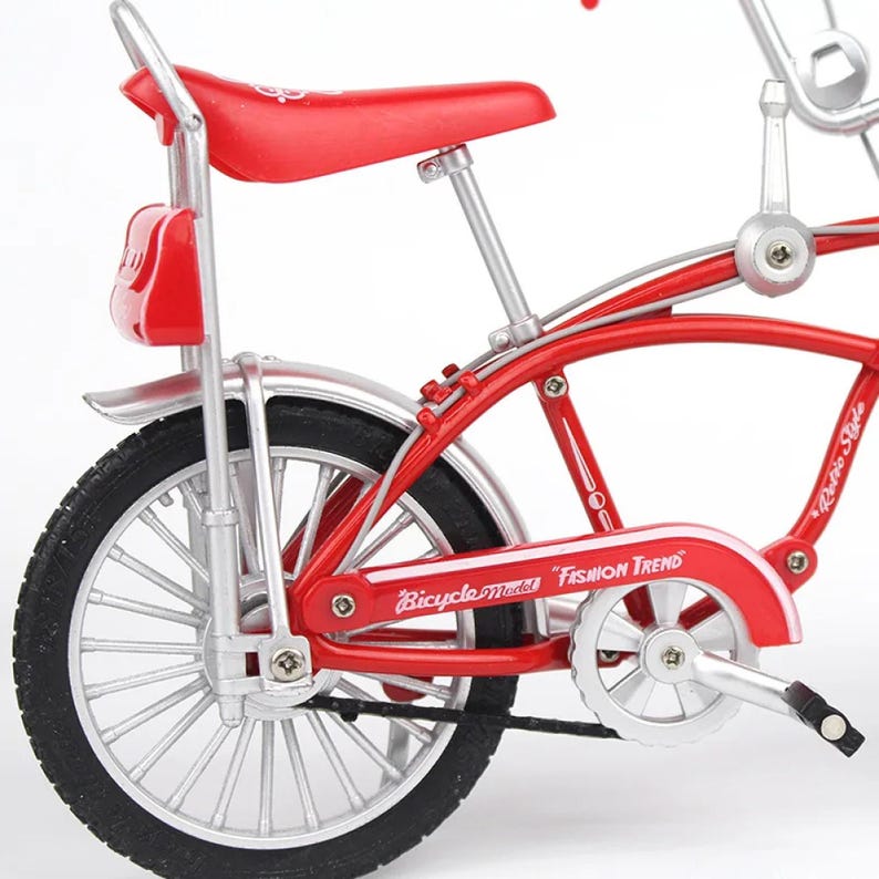 May include: A miniature red bicycle model with silver accents. The bike features a red seat, fenders, and frame, with black tires and a rear carrier. The words "Bicycle Model Fashion Trend" are visible on the frame.