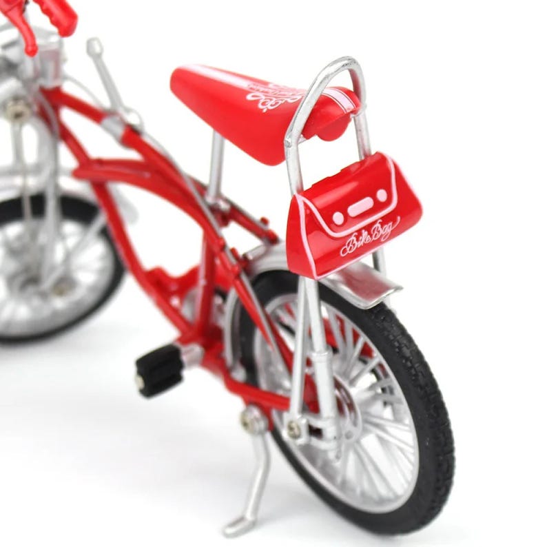 May include: A miniature red bicycle with silver accents. The bike features a red seat, handlebars, and frame. A small red bag with the text "Babe Bag" is attached to the back. The tires are black.