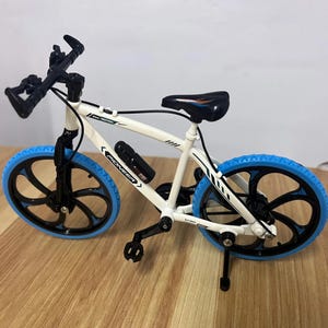 Op de afbeelding: Een witte miniatuurfiets met zwarte accenten en blauwe banden. De fiets heeft een zwart zadel, stuur en een bidonhouder. De wielen hebben een uniek spaakontwerp en de banden zijn helderblauw.