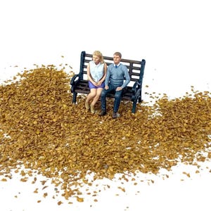 May include: Miniature figures of a man and woman seated on a black park bench. The woman wears a white top and purple skirt, the man a blue sweater and jeans. The scene is set amidst a bed of brown, leaf-like material.