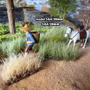 Może przedstawiać: Miniaturowa scena przedstawiająca postać niosącą kosz, białego konia z siodłem i wysoką trawę. Postać ma na sobie słomkowy kapelusz i niebieską koszulę. Widoczny jest tekst "model 1:64/28mm". Scena rozgrywa się na polnej ścieżce.