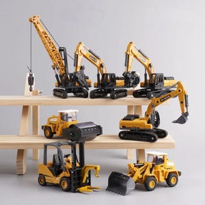 May include: A selection of yellow and black toy construction vehicles, including a crane, excavators, a road roller, a forklift, and a front-end loader. The toys are displayed on a light-coloured wooden shelf against a neutral background.