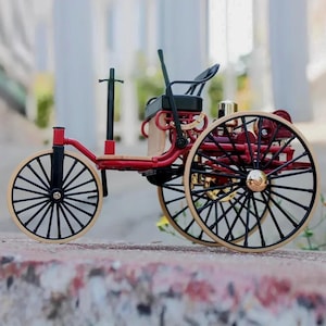 Può includere: Modello in miniatura di auto d'epoca con telaio rosso, ruote nere e dettagli dorati. Le ruote hanno raggi neri e cerchi beige. L'auto ha un sedile nero e un vano motore in legno. Un oggetto decorativo.
