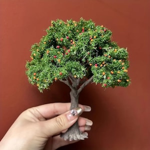 May include: A small model tree featuring a brown trunk and a dense, green crown adorned with tiny red and orange fruits. The tree is set against a plain, reddish-brown backdrop, highlighting its intricate design.