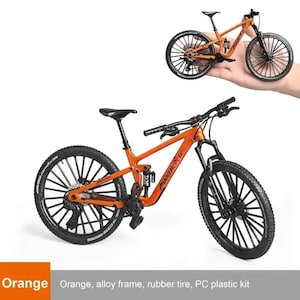 Op de afbeelding: Een oranje model mountainbike met zwarte banden en onderdelen. De fiets heeft een lichtmetalen frame en is een miniatuur replica. De afbeelding toont ook een kleinere versie van de fiets die in een hand wordt gehouden.