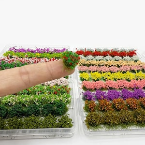 May include: Miniature, colorful model landscaping plants in clear plastic containers. The plants are arranged in rows and include various shades of green, red, pink, yellow, purple, and white. A small cluster of the plants is held between a finger and thumb.