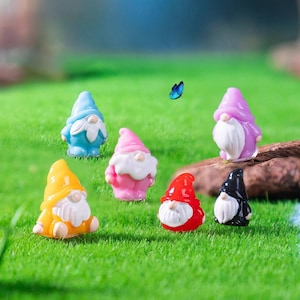 May include: A collection of miniature garden gnomes in various colors, including blue, pink, yellow, red, purple, and black. Each gnome has a white beard and a pointed hat. A blue butterfly flutters nearby, set against a backdrop of green grass.